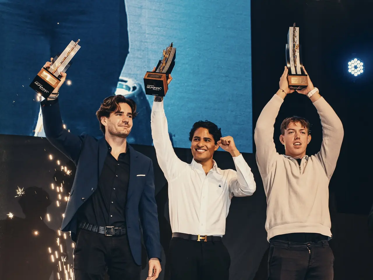 L'immagine mostra tre uomini in piedi su un palcoscenico, ciascuno con un trofeo in alto in segno...