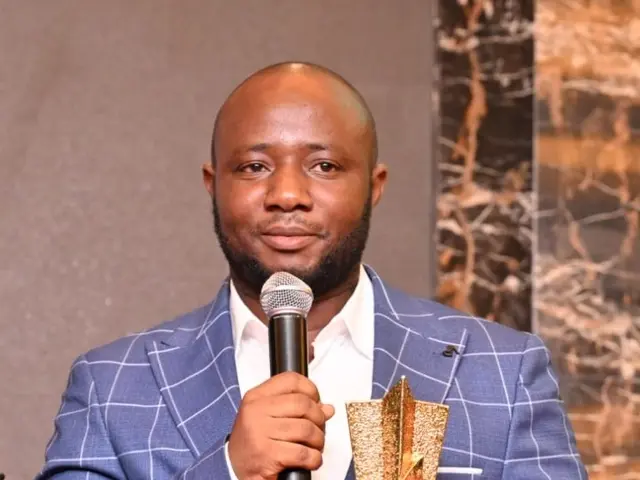 L'immagine mostra un uomo in giacca blu con un microfono e una coppa, con un muro sullo sfondo....